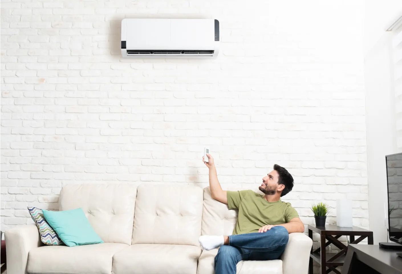 Relaxed man on a beige sofa pointing a remote at a ductless mini split air conditioner mounted on a white brick wall, enjoying efficient cooling in a modern living room.
