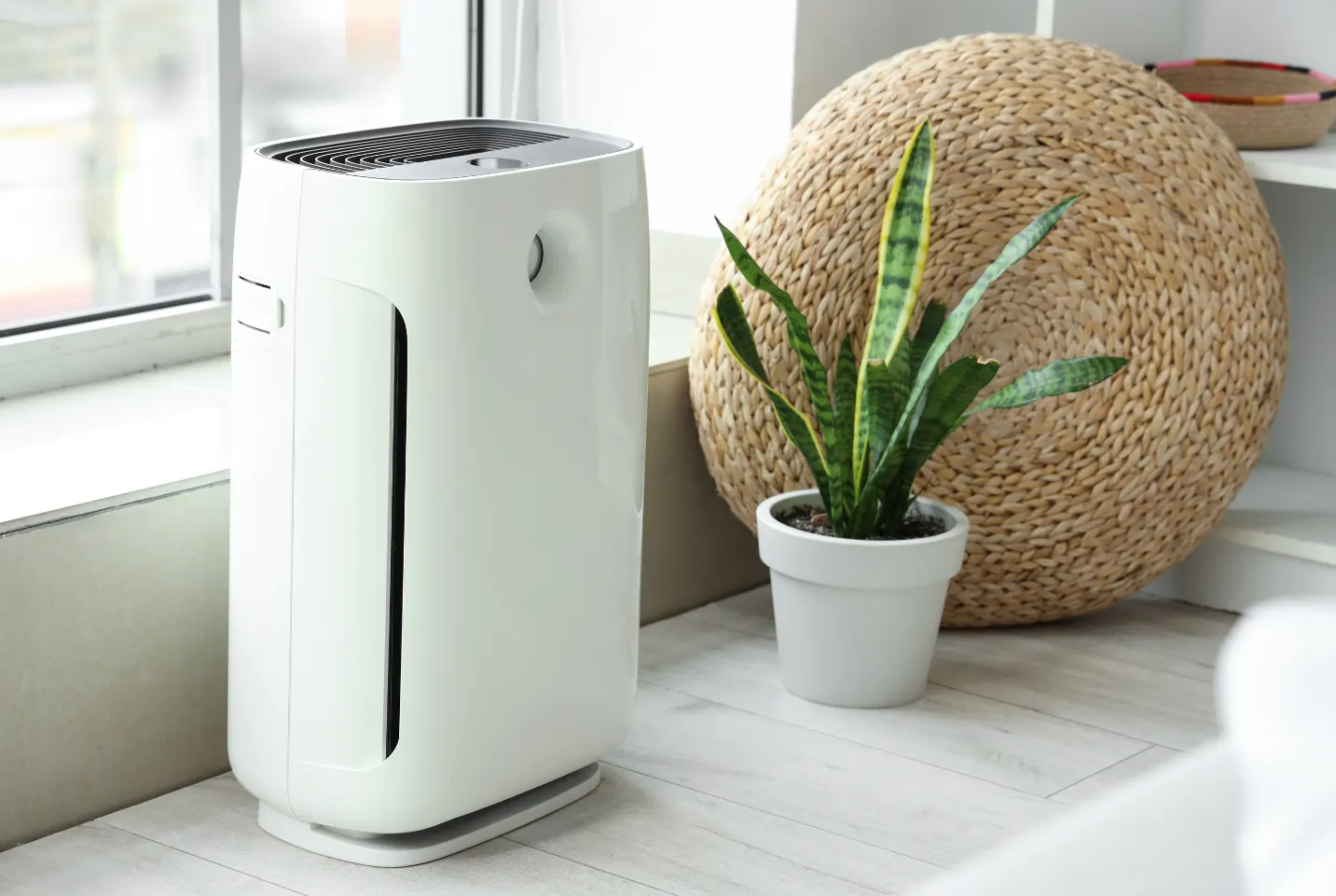 A sleek white air purifier placed next to a potted plant and a woven decorative mat by a window, enhancing indoor air quality in a cozy, sunlit home.