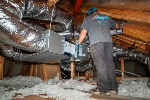 A technician in an attic working on HVAC ductwork and ventilation, surrounded by reflective foil insulation and loose-fill insulation on the attic floor. The technician is adjusting equipment with various cables and ducts extending across the attic space.